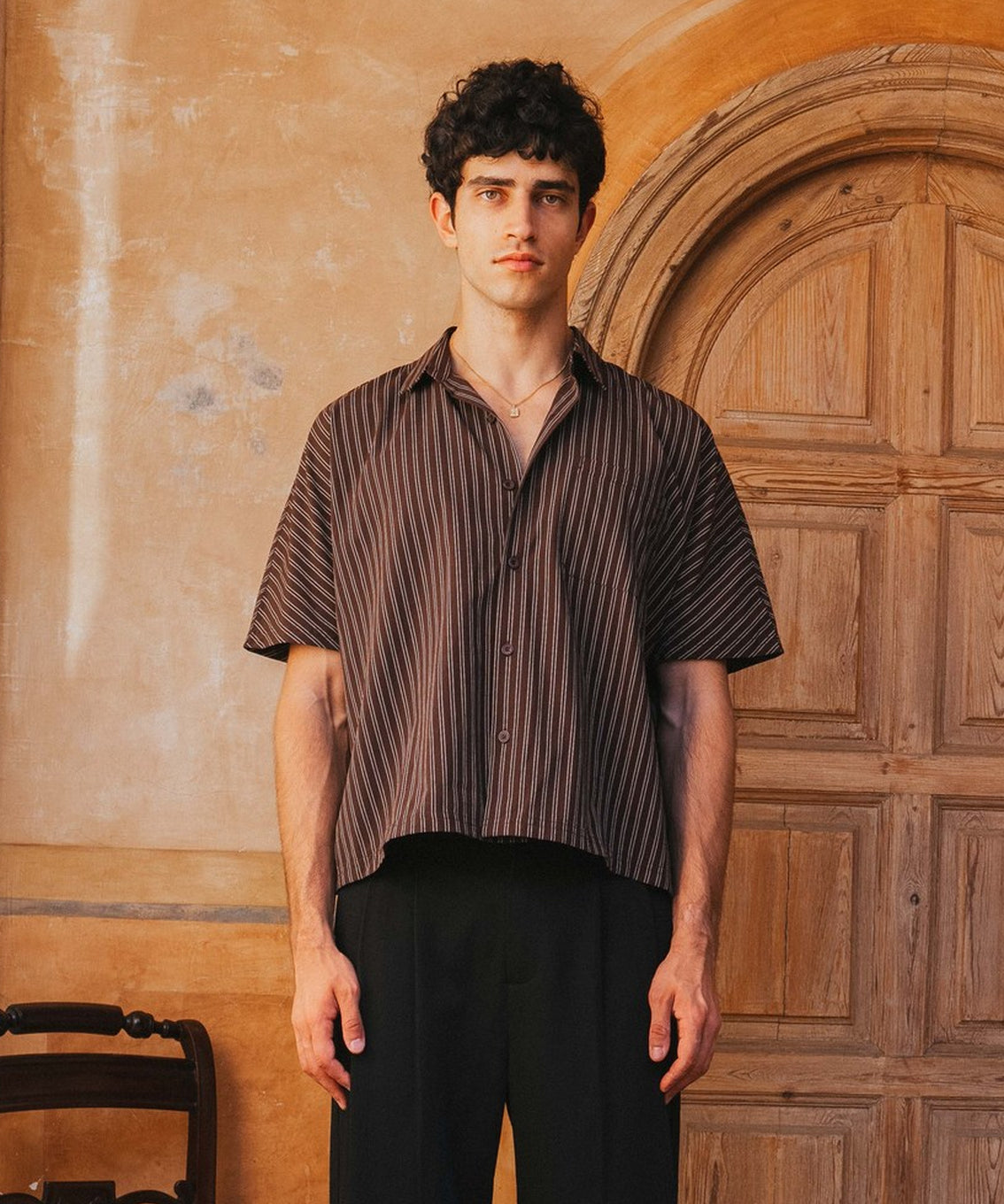 Man wearing a striped shirt and black pants standing in front of a wooden door.