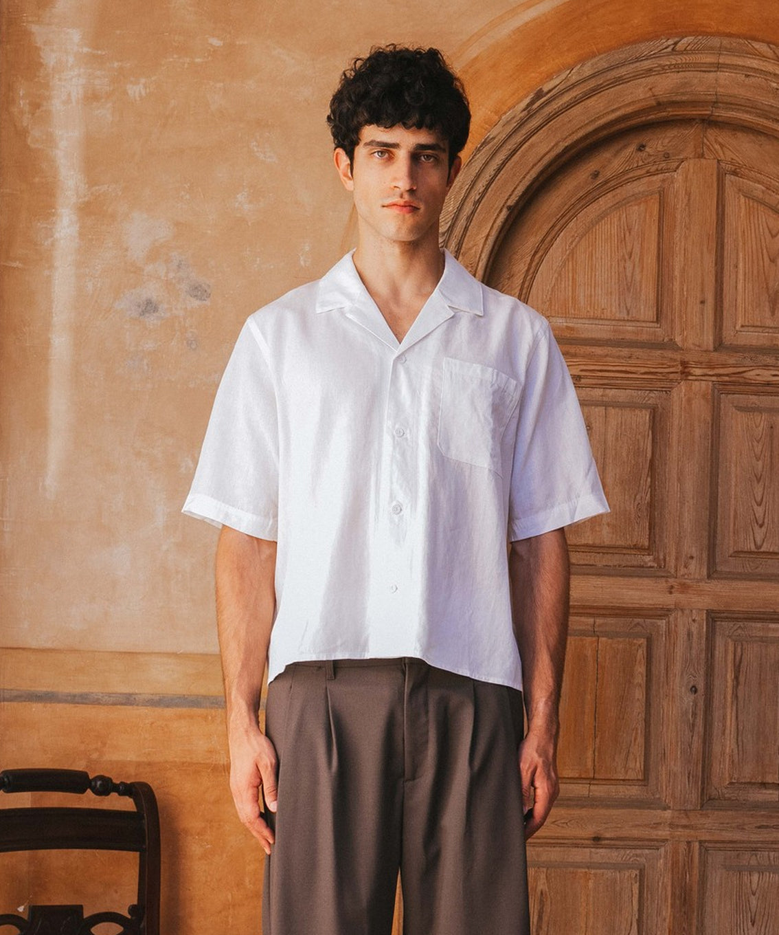Man wearing a white shirt and brown pants standing in front of a wooden door.