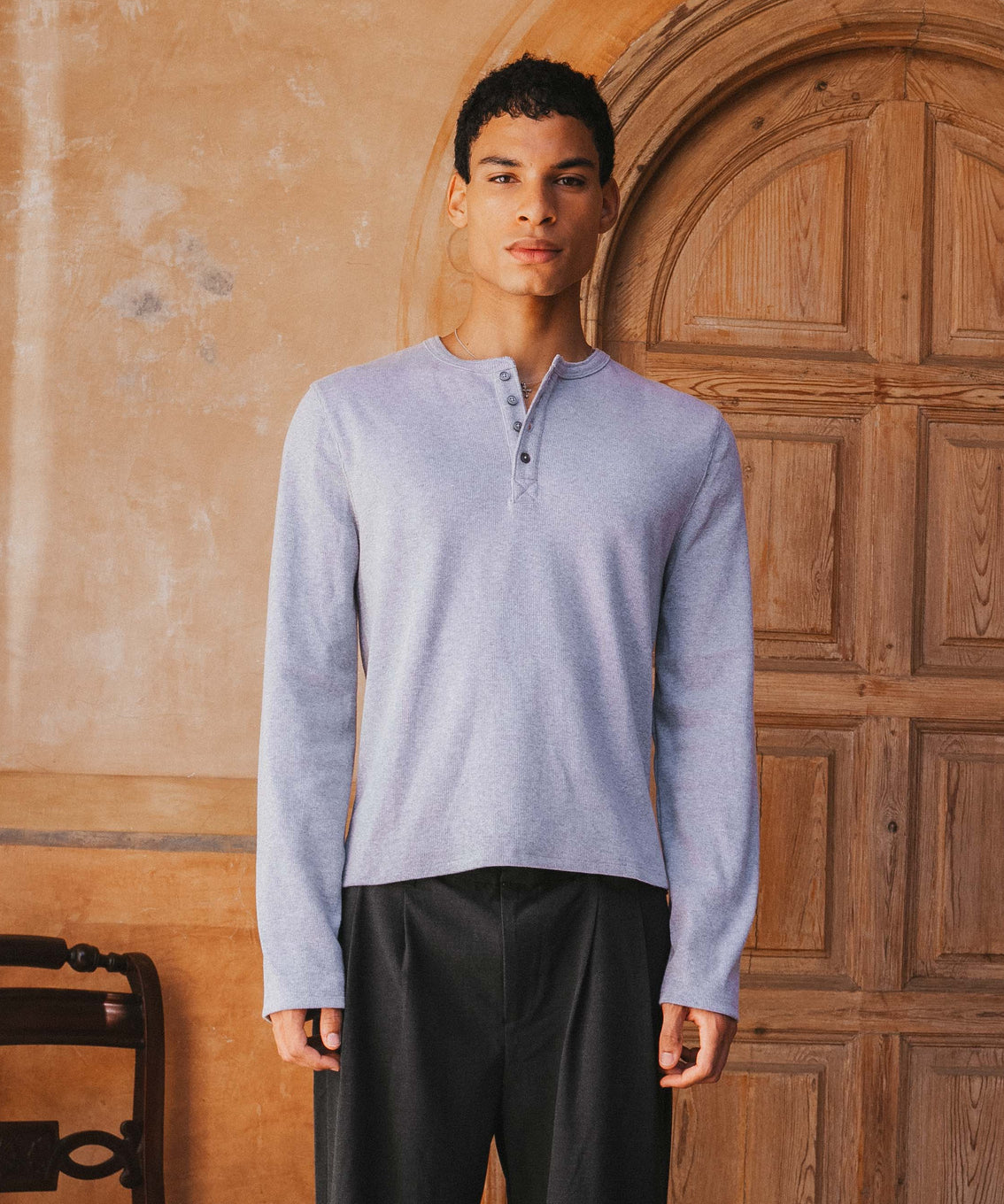 Man wearing a light blue long-sleeve shirt and dark pants standing in front of a wooden door.