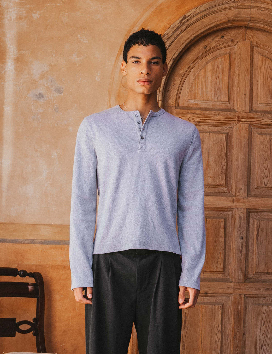 Man wearing a light blue long-sleeve shirt and dark pants standing in front of a wooden door.
