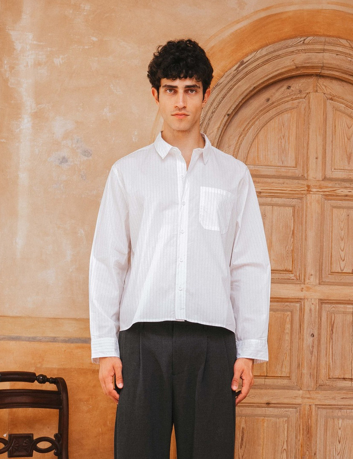 Man wearing a white shirt and black pants standing in front of a wooden door.