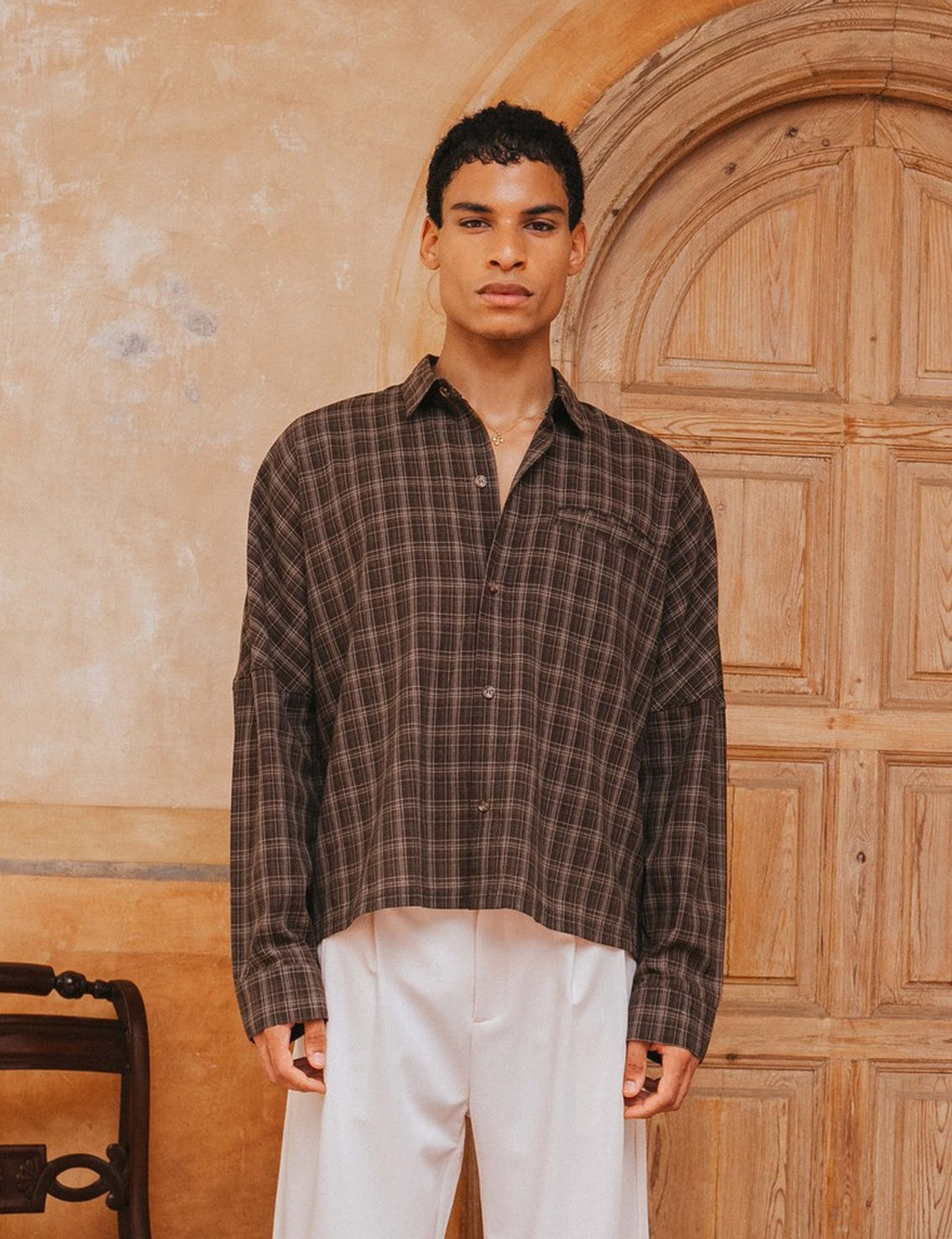 Man wearing a plaid shirt and white pants standing in front of a wooden door.