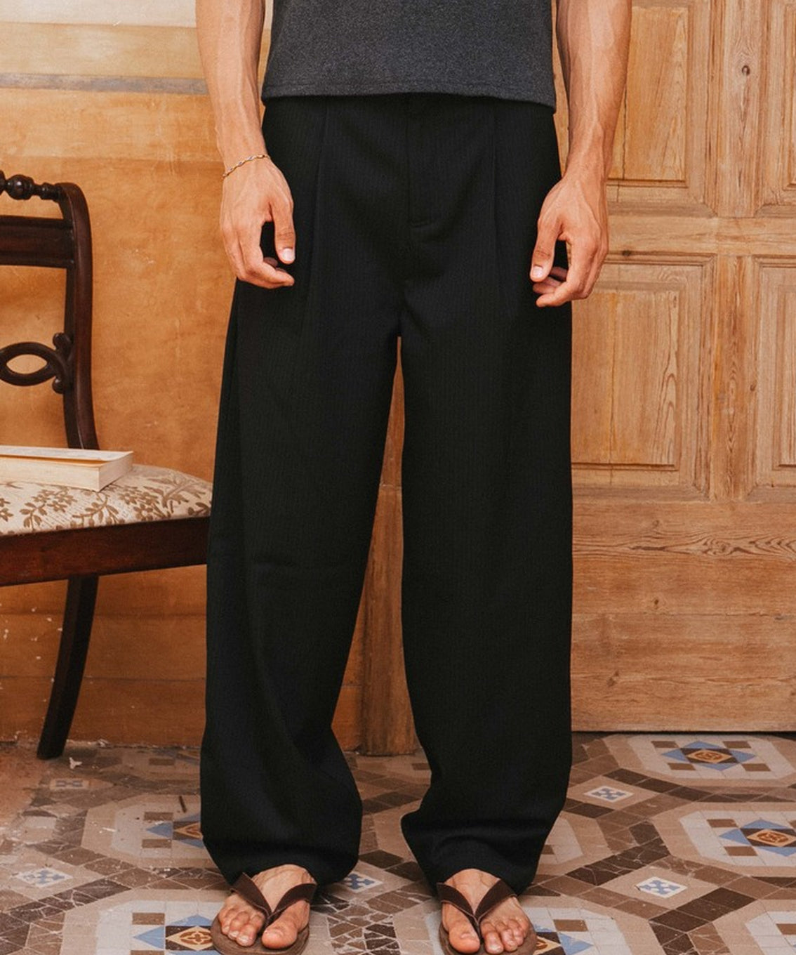 Person wearing black pants and a gray tank top standing in a room with wooden paneling and patterned floor.